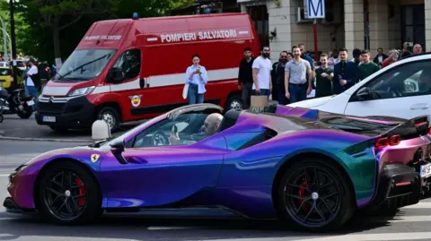 AFP via Getty Images Andrew Tate driving a purple, open-top Ferrari with a crowd in the background