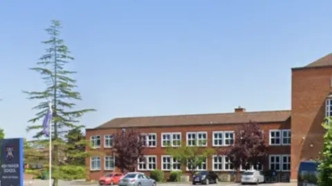 a long red brick building with a flag and sign for Ash Manor School at the entrance, surrounded by trees and a car park