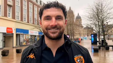 Jasmine Lowe/BBC News A man smiling at the camera with brown curly hair. He's wearing a black jacket with a Hull City crest on and a navy top with his top button open. He's stood in the centre of Hull where it has been raining.