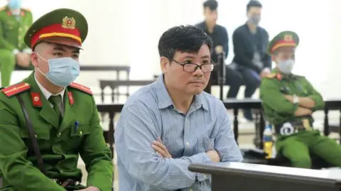 Getty Images A man with black hair wearing glasses and a light blue striped shirt sits with his arms folded across his chest. He is flanked by Vietnam security officials wearing uniforms and face masks