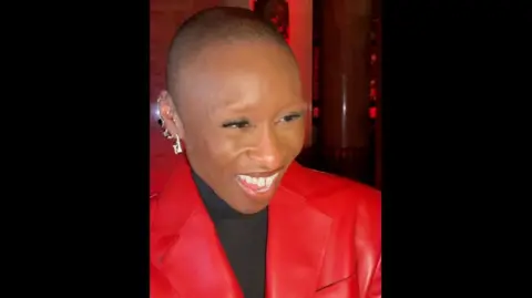 Cynthia Erivo is smiling, wearing a red jacket with a black turtleneck underneath.