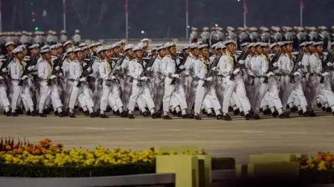 Jonathan Head/ BBC Soldiers in white uniforms holding weapons and marching in step at this year's armed forces parade.