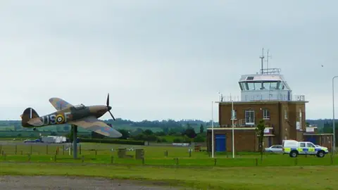 Geograph/Rod Allday A small fighter aircraft is on a plinth outside a watchtower at North Weald Airfield. There are grass lawns and some hills in the background.
