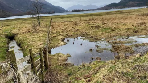 The Highland Council A boggy and grassy area of land near Glencoe Village with wooden fencing surrounding it. There is a loch and mountains in the background.