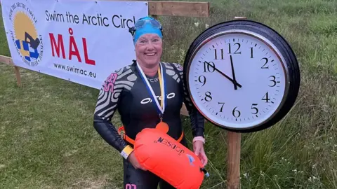 A female swimmer in a wetsuit holding a float and wearing a swimming cap and medals around her neck. She is stood beside a clock with the time reading 11:50 and behind her is a sign reading "swim the arctic circle"