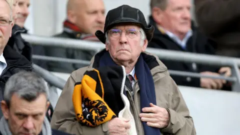 Sir Tom Courtenay, a man in his 80s, with grey hair, is standing in a football terrace with fans around him. he is wearing a black deer-stalker style hat and carrying a black and amber Hull City scarf.