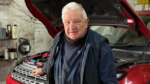 Rowland Neill had grey hair and is wearing a blue jumper and blue jacket. He is standing inside a garage at the front of a red car which has its bonnet up and is looking at the camera.
