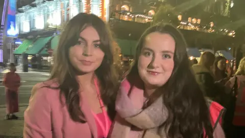 two women are standing in front of the City Hall. The women on the left have brown hair, are wearing a pink blazer with a pink top underneath. The other wears a pink and gray scarf with a pink hi-viz jacket, she has brown hair