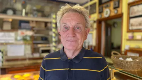 A customer with grey hair and a striped polo top standing in the bakery with pastries behind him