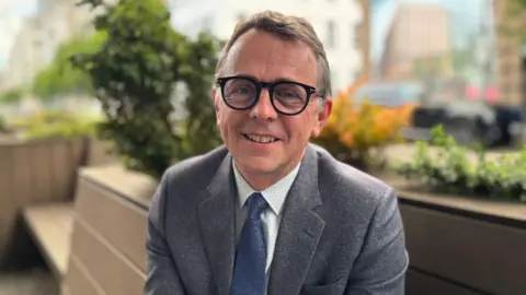 Professor Robbie McDonald. He has short brown hair, wearing glasses, a grey blazer, white shirt and blue tie.