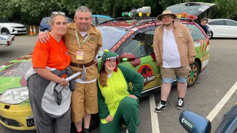 Justin Dealey/BBC Four people in dinosaur and Jurassic Park fancy dress costumes stand in front of a Jurassic Park themed estate car.