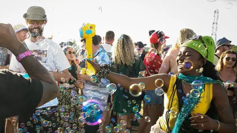 Getty Images A woman is dancing in bubbles. She is wearing a green hat and yellow top. She is standing in a crowd of people.