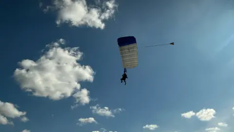 Emma Baugh/BBC Dan Perkins parachuting with a white and red parachute, with blue sky and white clouds