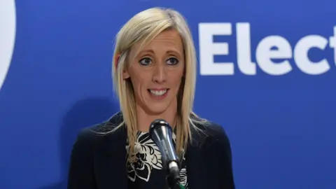 PA Media Carla Lockhart, with long blonde hair, wearing a black and white top and a black blazer. She is standing in front of a blue wall. 