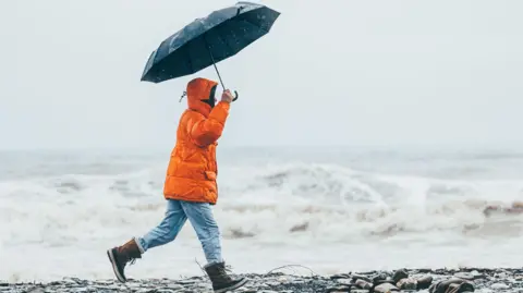 Getty Images A man wearing a bright orange coat, blue denim jeans and brown boots walks along the seashore. He is holding a navy umbrella.
