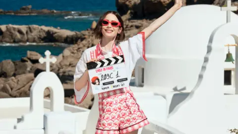 Lilly Collins stands with a clapperboard in a white t-shirt and red and white shorts. She extends one arm and wears red sunglasses. In the background is a white church and the sea of Greece.