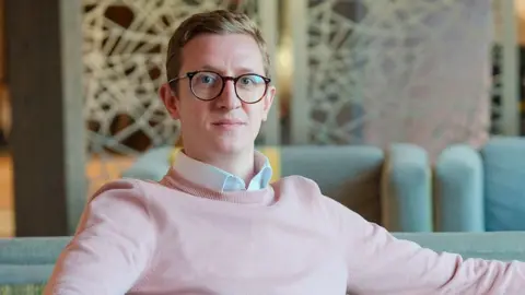 A man wearing glasses and a light pink jumper while seated on a grey sofa in the foreground. One arm rests along the back of the sofa and the other on the armrest. Additional sofas and a patterned room divider are visible in the background of the indoor lounge area.
