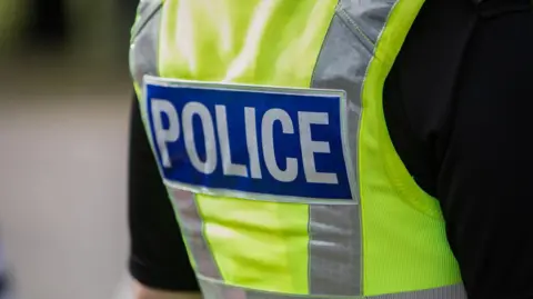 The back of a police officer's yellow high visibility vest with the word POLICE emblazened across it on a blue rectangle.