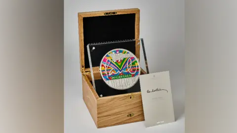 Royal Mint A silver coin which has Paul McCartney's signature on, along with a colourful striped background. It is presented in a wooden box.