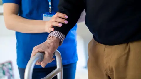 Close Up Of Senior Man With Hands On Walking Frame Being Helped By Care Worker.