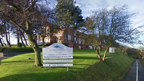 A red brick Victorian school building with trees and grass in front. A sign says Scarborough College: Independent Education for Boys and Girls Aged 3-18 Years Old