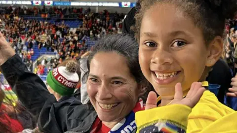 Marisa Teodósio Photograph of Maria Teodosio (right), pictured with her young daughter Yasmin (left) in a football stadium stand. She smiles at the camera showing her teeth and with her hand in the air.  