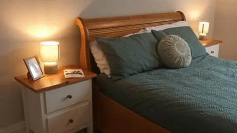 Missing Link A bedroom in the mental house crisis house with a green bedspread, two small chests of draws, pillows, lamps and a cushion.