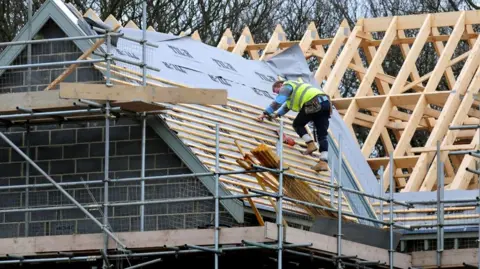 A man in a hi-vis vest works on a rood. He is placing wooden beams on the roof. There is scaffold around the rood and a wooden roof frame in the background.