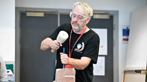 Rob MacDonald working on a stone sculpture. He is wearing a black t-shirt, protective glasses and a red lanyard. He has white hair and beard. There is a grey door behind him. 