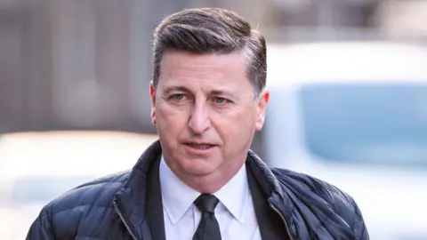 A man with dark hair, wearing a dark jacket, white shirt and dark tie, in a close-up shot 