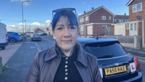 A woman with dark hair, a dark top and dark jacket with dark glasses in a street with a dark car behind her with L plates on the back