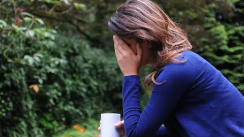 A woman wearing a blue long-sleeve shirt sits with her left-hand on her face, while her right hand holds a white mug. She is sat in a garden and from her body language she looks in distress. 