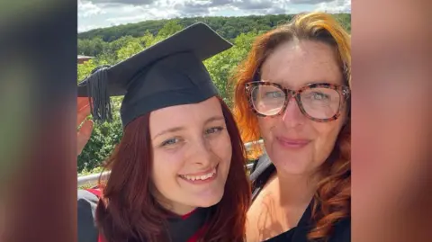 Ashley Cadet Daisy Cadet and her mother Ashley are smiling. Daisy wears a graduation gown and hat, and has red hair. Her mother Ashley is also smiling at the camera, has red hair and is wearing a black shirt. 