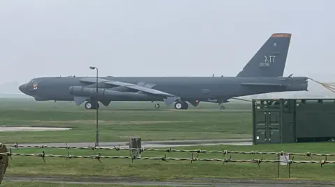 A large black B52 military bomber, a long sleek black plane, coming down the runway at Fairford air base. It is a misty day and there is barbed wire in the foreground. 