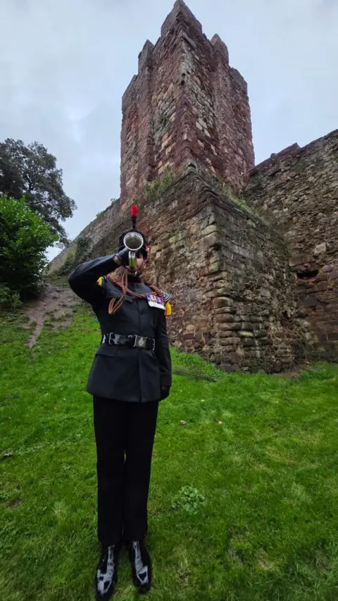 Johnny Rutherford/BBC A man in a military uniform plays the bugle. He is standing up straight. Behind him is a stone structure.