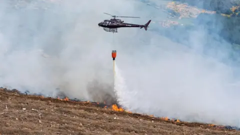 A helicopter waterbombs flames in a wildfire on a barren hillside. Water pours from a large red bucket under the helicopter.