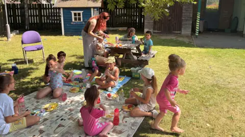 Children running or sitting to eat under the shade of a tree at The Parenting Network outside space. An adult woman is seen helping one of them at a table. It is sunny.