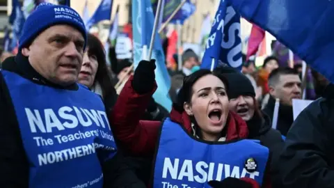 Charles McQuillan A picture of an NASUWT picket line, in the forefront are a man and woman wearing blue NASUWT bibs, with the woman waving a NASUWT flag and the man wearing a union hat. They are surrounded by other union members, many of them waving flags 