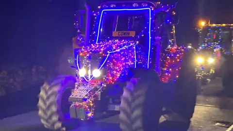 A tractor covered in strings of blue and red lights, with another following on the road.