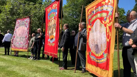 Tom Ingall/BBC Three colourful banners, each about two metres square, are held up by men in suits