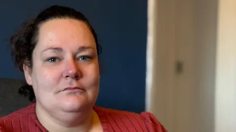 A woman with short dark hair looks at the camera. The photo is close-up, showing her face and shoulders. She wears an red top and the background is blurred.