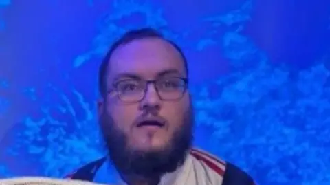Nathan Major-Kershaw Man in glasses with dark hair and dark beard wearing a red, white and blue top with a bright blue background