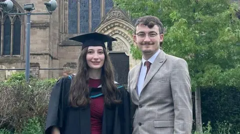 A woman in a dark red dress wears a black graduation gown and cap; she has brown hair and is smiling at the camera. A man with brown hair and glasses is standing next to her in a tweed jacket 