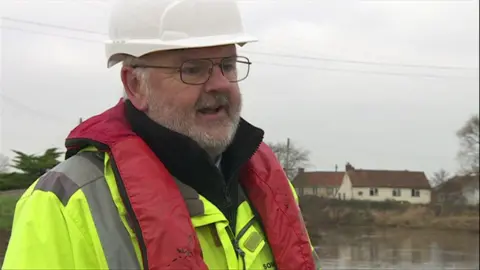 Iain is in the middle of the shot looking out to the right of the camera. He is wearing glasses and a hard hat, as well as a hi-vis coat, red buoyancy aid and black jumper. Behind him is the river and some houses are in the distance.