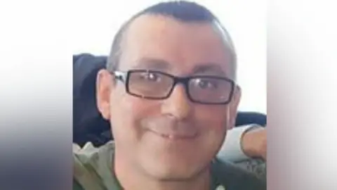 West Yorkshire Police A man with rectangular black glasses smiles at the camera. He is wearing a green hoodie.