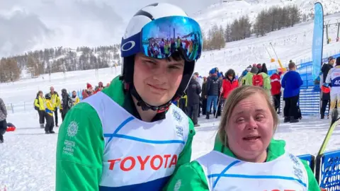 BBC Caolán McConville left and Lucy Best right. Both are smiling at the camera. Caolán is wearing a ski helmet. Lucy has blonde hair . They are wearing green jackets with a white vest on top. They are standing at the ski slopes in Italy. 