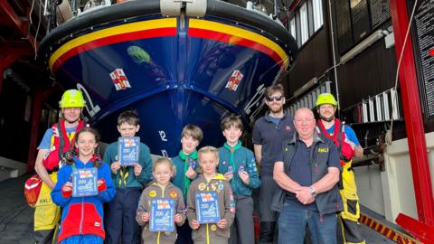 A group of children with RNLI volunteers smiling with badges in front of a lifeboat