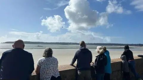 Emily Bell/BBC Seven people, including a child have their backs to the camera as they look out over St Aubin's Bay at a aircraft approaching with a trail of white cloud in its wake.