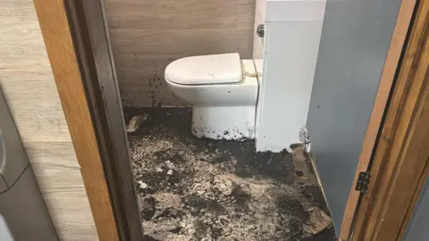 Handout Dark ashes on the floor of a small cubicle, with a white toilet.