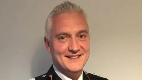 Ben Brook, West Midlands Fire Service, in his firefighter uniform in front of a white wall 